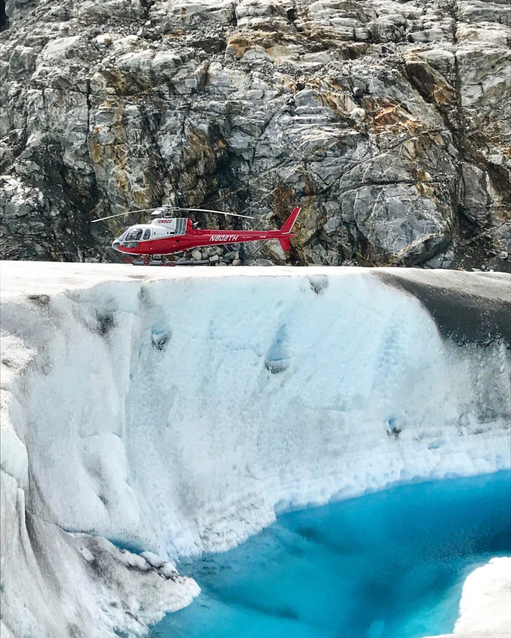 A helicopter sets down on a glacier near a blue pool of water.