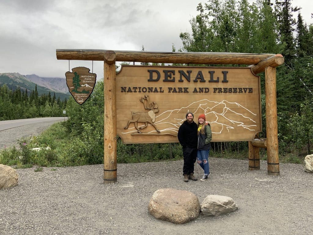 A couple stands in front of the Denali National Park sign in Denali Alaska.